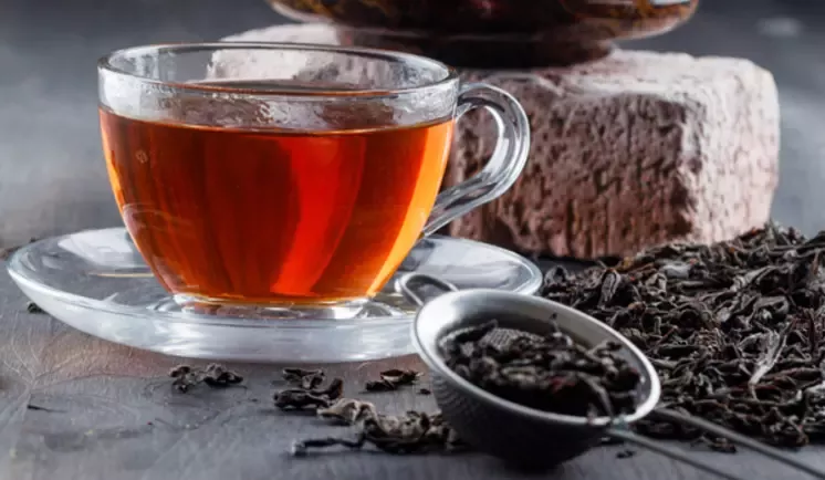 فنجان شیشه‌ای چای روی نعلبکی، کنار برگ‌های خشک چای و قاشق روی میز چوبی